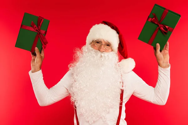 Santa Claus en sombrero sosteniendo regalos y mirando a la cámara aislada en rojo - foto de stock