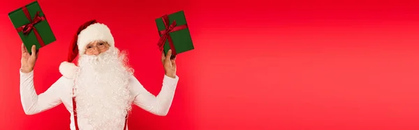Santa claus in eyeglasses and hat holding presents isolated on red with copy space, banner — Stock Photo