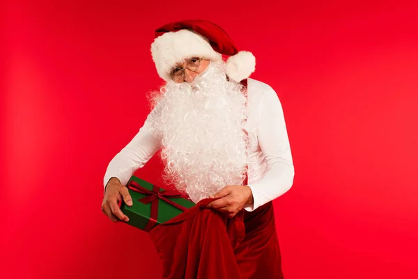 Father christmas in hat putting present in sack isolated on red — Stock Photo
