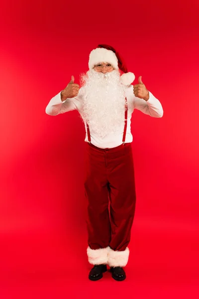 Toute la longueur de père barbu Noël montrant pouces vers le haut sur fond rouge — Photo de stock