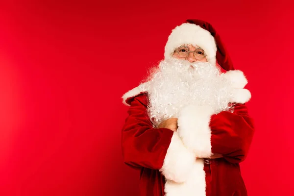 Бородатий Санта Клаус схрещує руки і дивиться на камеру на червоному тлі — стокове фото