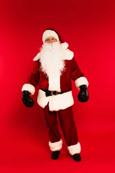 Full length of santa claus in costume and boxing gloves standing on red background — Stock Photo