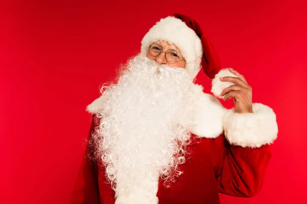 Babbo Natale barbuto in occhiali toccante cappello isolato su rosso — Foto stock