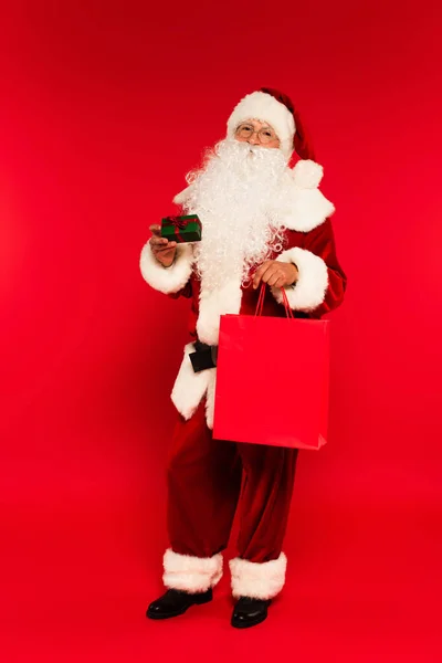 Full length of santa claus holding shopping bag and small gift box with bow on red background — Stock Photo