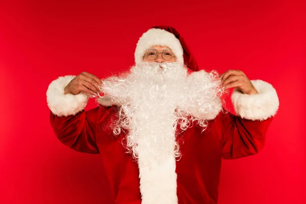 Père Noël dans des lunettes de vue touchant la barbe sur fond rouge — Photo de stock