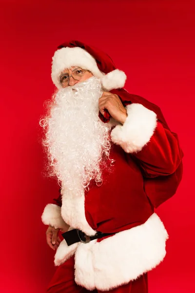 Санта-Клаус в очках держит мешок на красном фоне — стоковое фото