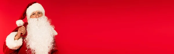Santa Claude dans des lunettes tenant sac et regardant la caméra isolée sur rouge avec espace de copie, bannière — Photo de stock