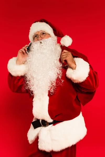 Papai Noel barbudo segurando saco e falando no smartphone no fundo vermelho — Fotografia de Stock