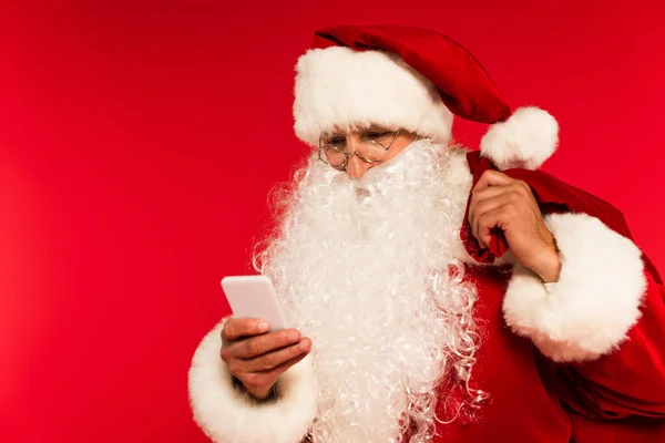 Санта-Клаус в очках держа мешок и используя смартфон изолирован на красный — стоковое фото