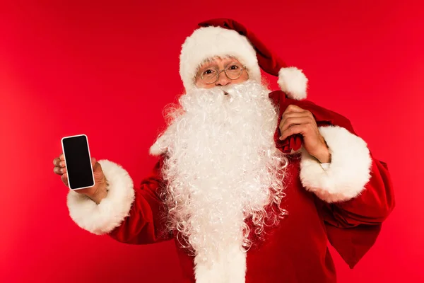 Weihnachtsmann mit Sack hält Smartphone mit leerem Bildschirm auf rotem Hintergrund — Stockfoto