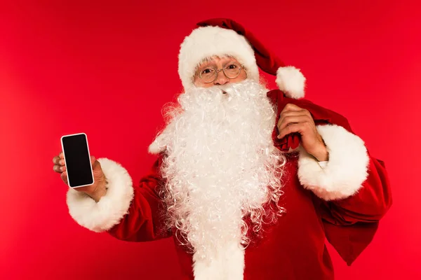 Father chistmas with sack holding smartphone with blank screen on red background — Stock Photo
