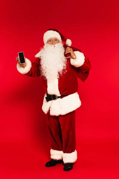 Santa Claus en traje sosteniendo saco y teléfono celular con pantalla en blanco sobre fondo rojo - foto de stock