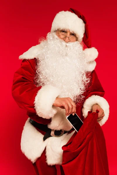 Santa Claus en anteojos poniendo smartphone en saco aislado en rojo - foto de stock
