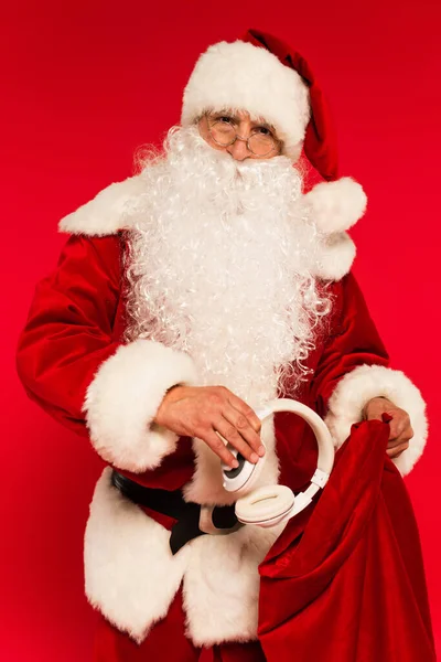 Santa claus dans les lunettes et le chapeau mettre des écouteurs dans le sac isolé sur rouge — Photo de stock