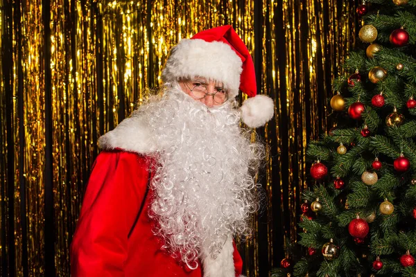 Санта-Клаус в костюмах і окулярах дивиться на камеру біля ялинки і золотої мішури — стокове фото