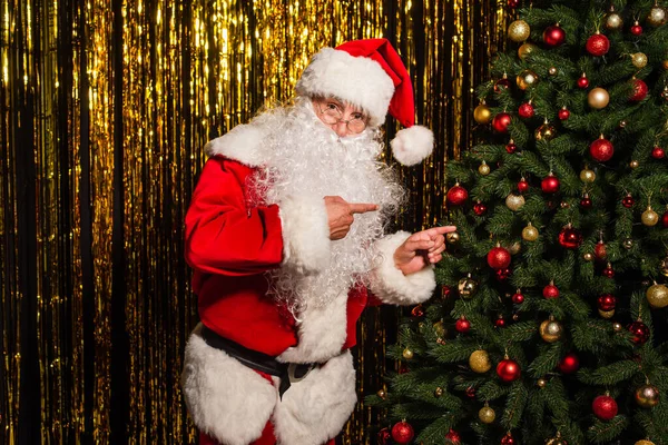 Santa claus pointing with fingers at decorated christmas tree near tinsel — Stock Photo