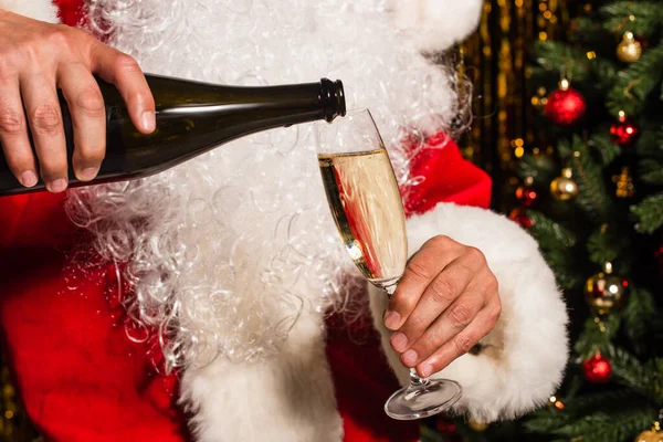 Vista cortada de Papai Noel barbudo derramando champanhe perto de árvore de Natal borrada — Fotografia de Stock