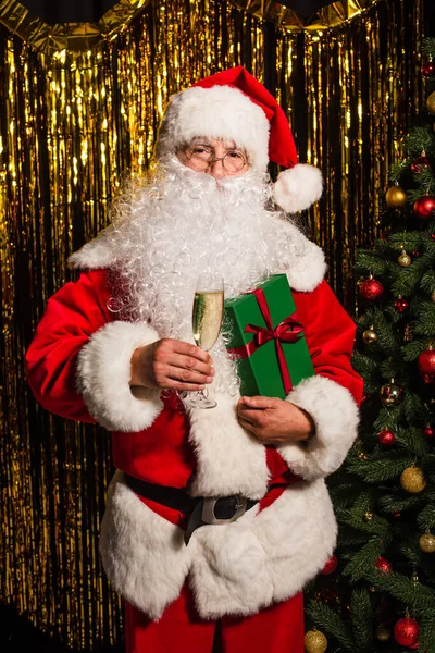 Santa claus holding glass of champagne and gift near christmas tree and tinsel — Stock Photo