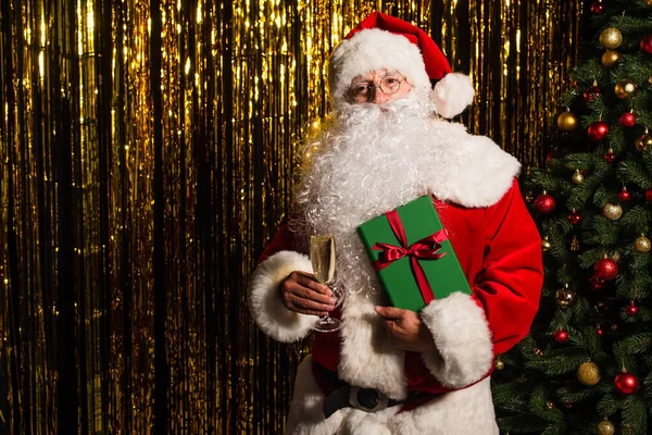 Santa Claus sosteniendo presente y champán cerca de árbol de Navidad y oropel - foto de stock