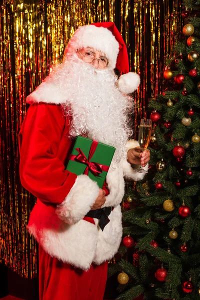 Санта-Клаус в очках с шампанским и подарками возле рождественской елки и мишуры — стоковое фото