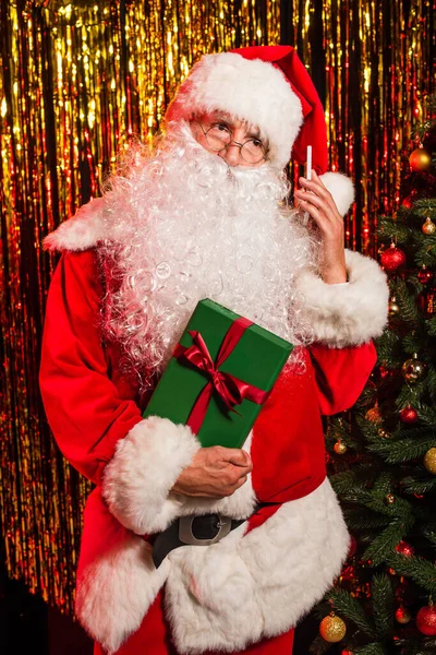 Santa claus tenant smartphone et cadeau près de l'arbre de Noël décoré et tinsel — Photo de stock