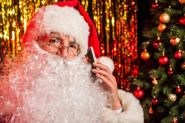 Bearded santa claus in eyeglasses talking on smartphone near blurred christmas tree and tinsel — Stock Photo