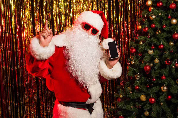 Santa Claus en gafas de sol sosteniendo smartphone con pantalla en blanco y apuntando con el dedo cerca del árbol de Navidad y el oropel - foto de stock