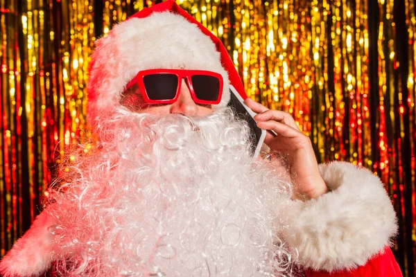 Bärtiger Weihnachtsmann mit Sonnenbrille spricht auf Smartphone neben verschwommenem Lametta — Stockfoto