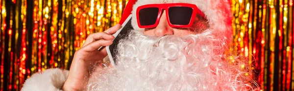 Bärtiger Weihnachtsmann mit Sonnenbrille, der neben verschwommenem Lametta auf dem Smartphone redet, Banner — Stockfoto