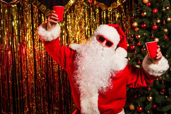 Santa Claus en gafas de sol con copas desechables cerca del árbol de Navidad y el oropel - foto de stock