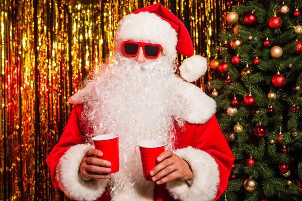Santa Claus en gafas de sol y sombrero sosteniendo copas de plástico cerca de árbol de Navidad y oropel - foto de stock