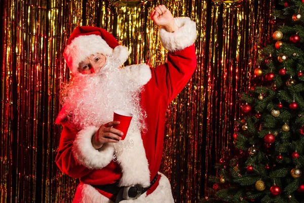 Возбужденный Санта Клаус держит пластиковую чашку возле рождественской елки и мишуры — стоковое фото