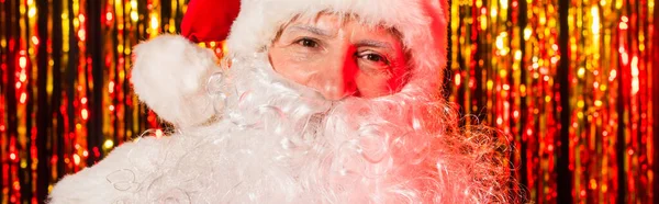 Portrait of santa claus in red hat looking at camera near blurred tinsel, banner — Stock Photo