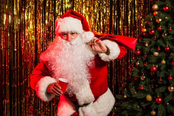 Papai Noel segurando copo de plástico e saco de compras perto de árvore de natal e ouropel — Fotografia de Stock