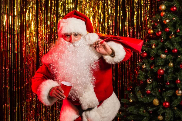 Babbo Natale in possesso di tazza di plastica e shopping bag vicino all'albero di Natale e fili di lame — Foto stock