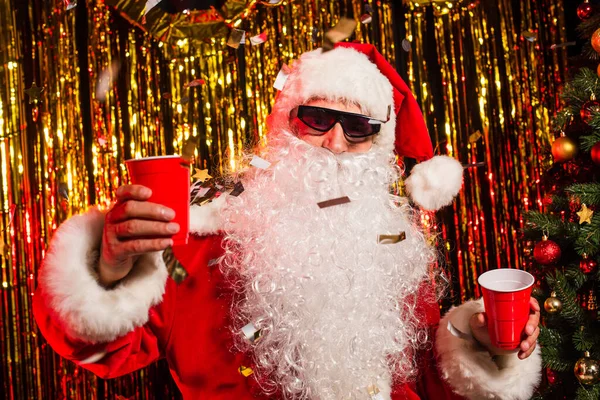 Papai Noel em óculos de sol segurando copos de plástico sob confete perto da árvore de natal — Fotografia de Stock