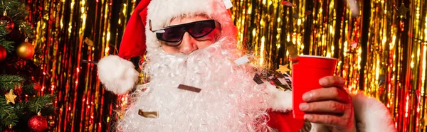 Santa Claude dans des lunettes de soleil tenant tasse en plastique sous confettis près de tinsel, bannière — Photo de stock