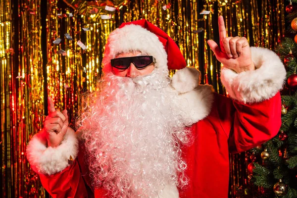 Father christmas in sunglasses pointing with fingers under confetti near festive tinsel — Stock Photo