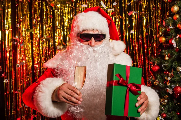 Santa claus in sunglasses holding champagne glass and present near christmas tree and tinsel — Stock Photo