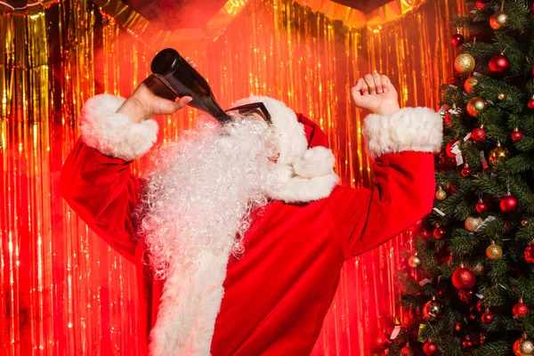 Papai Noel em óculos de sol bebendo champanhe de garrafa perto de árvore de natal e ouropel — Fotografia de Stock