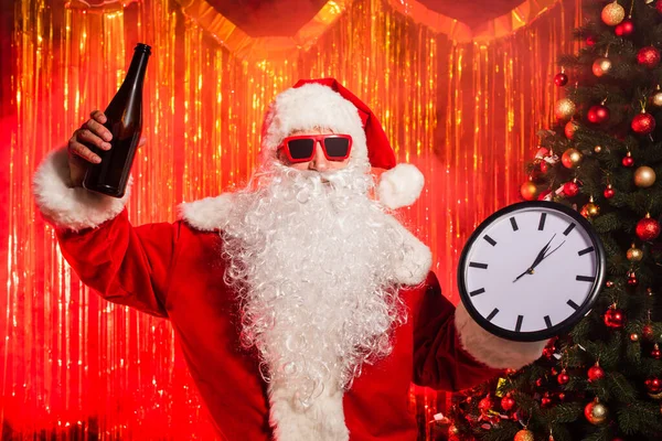 Santa Claude dans des lunettes de soleil tenant bouteille de champagne et horloge près de l'arbre de Noël — Photo de stock