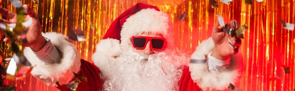 Santa Claus en gafas de sol y traje lanzando confeti durante la fiesta cerca de oropel, pancarta - foto de stock
