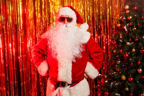Père Noël en lunettes de soleil et costume debout dans akimbo pose près de tinsel — Photo de stock