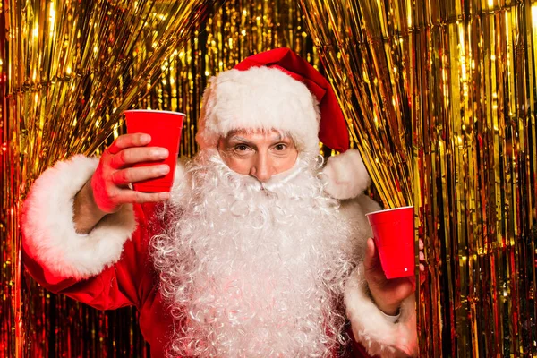 Papai Noel em traje segurando copos de plástico e olhando para a câmera perto de ouropel — Fotografia de Stock