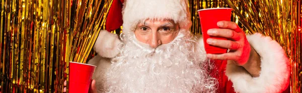 Barbuto padre Natale in possesso di bicchieri di plastica vicino fili di lame lucido, banner — Foto stock