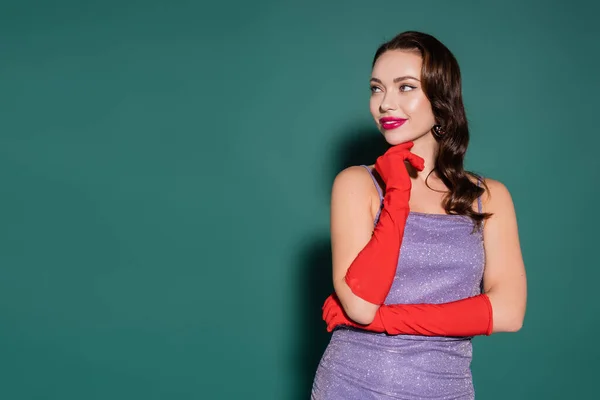 Mujer joven feliz en guantes rojos y vestido púrpura mirando hacia otro lado en verde - foto de stock