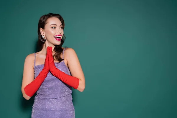 Mujer joven excitada en guantes rojos y vestido púrpura gesto mientras mira hacia otro lado en verde - foto de stock