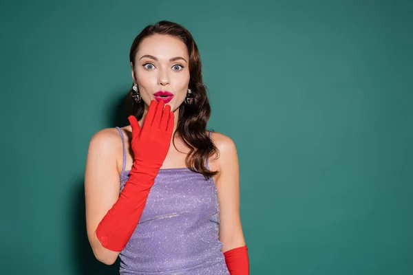 Shocked young woman in red gloves and purple dress covering opened mouth on green — Stock Photo