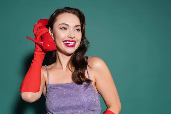 Cheerful young woman in red glove holding high heeled shoe near ear on green — Stock Photo