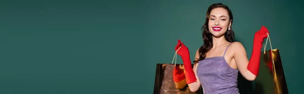 Happy young woman in red gloves and purple dress holding shopping bags on green, banner — Stock Photo